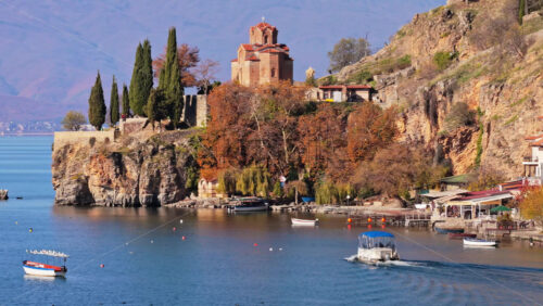 Video – Aerial drone view of the Church of St. John at Kaneo, surrounded by rocky cliffs, autumn trees, and small boats docked along the shoreline - Starpik Stock
