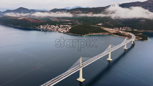 Video – Aerial drone view of the Peljesac Bridge near Komarna, Croatia, partially shrouded in low coastal fog with mountains in the background - Starpik Stock