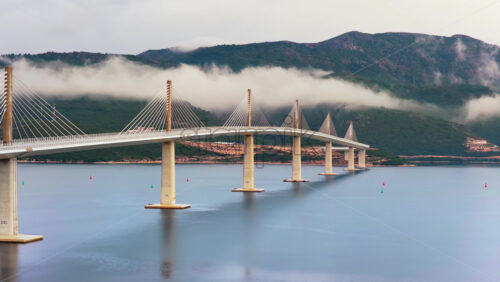 Video – Aerial drone view of the Peljesac Bridge near Komarna, Croatia, partially shrouded in low coastal fog with mountains in the background - Starpik Stock