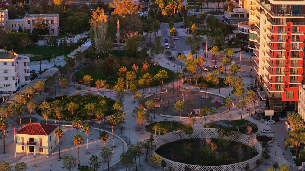Video – Aerial drone view of the golden hour lighting highlighting Vlora’s central urban park, featuring circular pathways, palm trees, playgrounds, and landscaped green spaces - Starpik Stock
