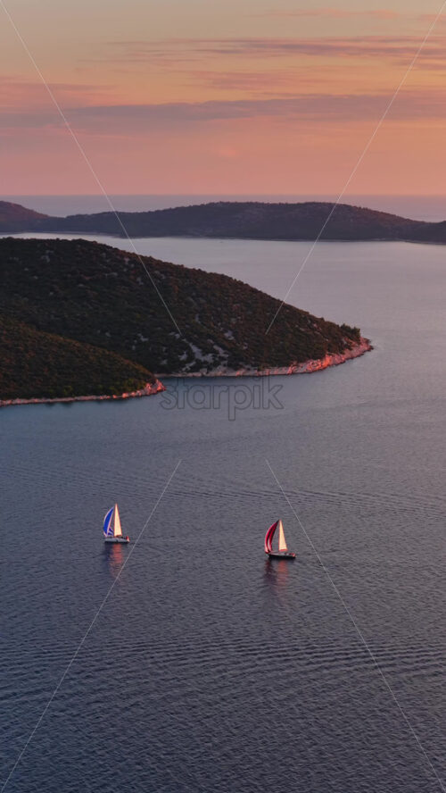 Video – Aerial drone view of two sailboats moving across calm water, with several green, forested islands in the background under pastel evening light. Vertical - Starpik Stock