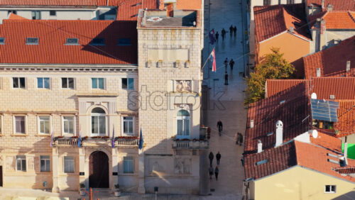 Video - Aerial drone view of an elegant stone building with flags and carved details, beside a narrow stone pedestrian street. Zadar, Croatia