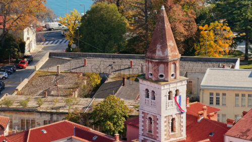 Video - Aerial drone view of a tall church tower displaying the Croatian flag, surrounded by autumn colored trees and rooftops. Zadar, Croatia