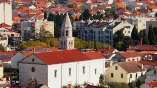 Video - Aerial drone view of a church with a tall stone bell tower and a red roof, surrounded by residential buildings and greenery in a densely built urban area. Biograd na Moru, Croatia