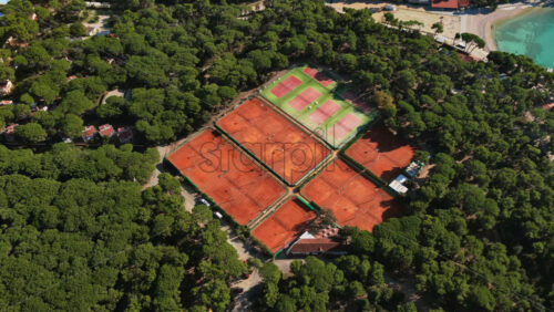 Video - Aerial drone view of a large tennis complex featuring several clay courts and green hard courts, surrounded by thick forest near the coastline