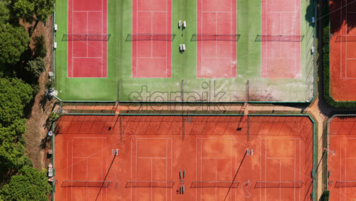 Video - Aerial drone view of a large tennis complex featuring several clay courts and green hard courts, surrounded by thick forest near the coastline