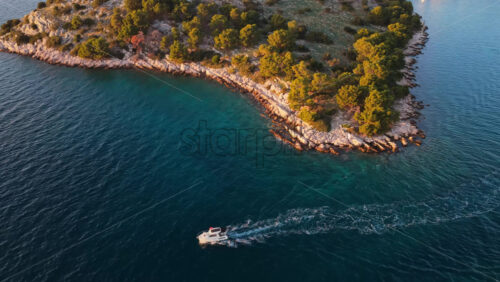 Video - Aerial drone view of a small motorboat moving along the rocky coastline of a forested island, leaving a white wake across the blue water