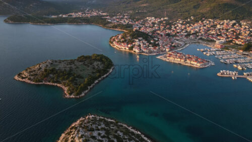 Video - Aerial drone view of rocky green islands separated by narrow channels of turquoise water, with a coastal town and marina visible in the background. Tribunj, Croatia