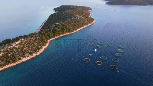 Video - Aerial drone view of a coastal island landscape near Tribunj, Croatia, highlighting clear blue Adriatic waters, rocky shorelines, dense pine forests, and an offshore circular fish farm