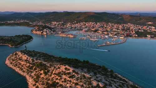 Video - Aerial drone view of rocky green islands separated by narrow channels of turquoise water, with a coastal town and marina visible in the background. Tribunj, Croatia