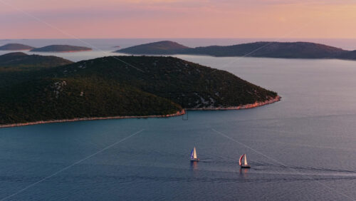 Video - Aerial drone view of two sailboats moving across calm water, with several green, forested islands in the background under pastel evening light. Adriatic sea, Croatia