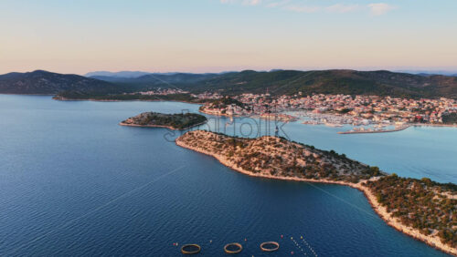 Video - Aerial drone view of a coastal island landscape near Tribunj, Croatia, highlighting clear blue Adriatic waters, rocky shorelines, dense pine forests, and an offshore circular fish farm