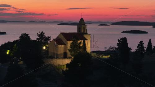 Video - Aerial drone view of a historic stone church stands on a hill facing a vivid pink orange sunset, with islands scattered across the horizon in Tribunj, Croatia