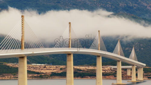 Video - Aerial drone view of the Peljesac Bridge near Komarna, Croatia, partially shrouded in low coastal fog with mountains in the background