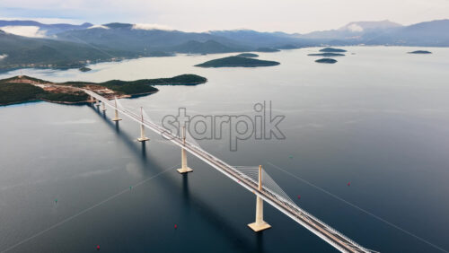 Video - Aerial drone view of the Peljesac Bridge near Komarna, Croatia, partially shrouded in low coastal fog with mountains in the background