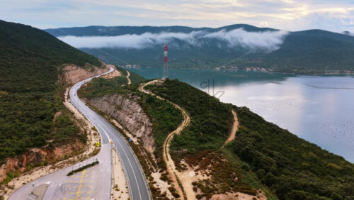 Video - Aerial drone view of the scenic coastal area around Peljesac Bridge, Croatia, featuring winding roads, dense green hills, oyster farms in the water, and fog rolling over the mountains