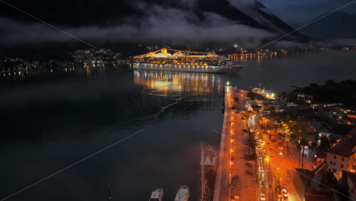 Video - Aerial drone view of a nighttime port scene with a brightly lit cruise ship docked near a waterfront lined with warm streetlights and moored yachts