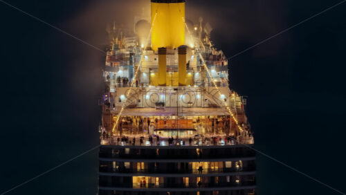 Video - Aerial drone view of a massive cruise ship, brightly lit with warm lights, as passengers enjoy its outdoor decks at night