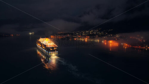 Video - Aerial drone view of a cruise ship moving slowly through a dark and misty bay at night, with faint lights of surrounding towns reflecting on the water near the Bay of Kotor
