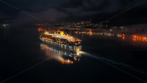 Video - Aerial drone view of a cruise ship moving slowly through a dark and misty bay at night, with faint lights of surrounding towns reflecting on the water near the Bay of Kotor