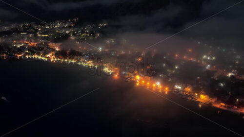 Video - Aerial drone view of the Bay of Kotor at night, with fog drifting across the landscape and a brightly lit cruise ship reflecting on the water. Coastlines glow with orange lights