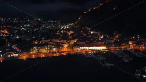 Video - Aerial drone view of the Bay of Kotor at night, with fog drifting across the landscape and a brightly lit cruise ship reflecting on the water. Coastlines glow with orange lights