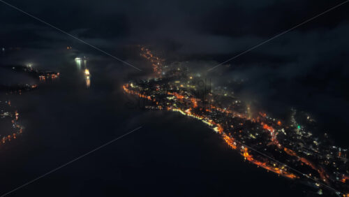 Video - Aerial drone view of the Bay of Kotor at night, with fog drifting across the landscape and a brightly lit cruise ship reflecting on the water. Coastlines glow with orange lights
