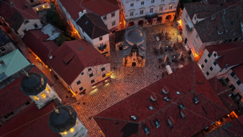 Video - Aerial drone view of a charming stone square in Kotor Old Town, centered around a historic chapel and surrounded by cafes and traditional buildings. Montenegro