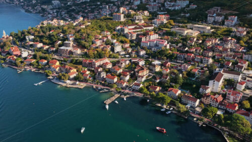 Video - Aerial drone view of a seaside neighborhood with red rooftops, tree lined streets, and small piers along the shoreline. Bay of Kotor, Montenegro