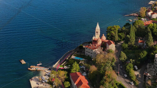 Video - Aerial drone view of a beautiful coastal church with a tall bell tower and terracotta roofs, surrounded by trees and blue sea. Bay of Kotor, Montenegro
