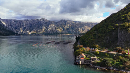 Video - Aerial drone view of a small lighthouse and shoreline road curving around a rocky hill, with dramatic mountain scenery across the bay. Bay of Kotor, Montenegro