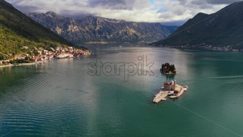 Video - Aerial drone view of Perast in the Bay of Kotor, showing Our Lady of the Rocks and Saint George Island centered on calm turquoise water, with the coastal village and dramatic mountains in the background