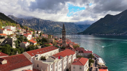 Video - Aerial drone view of the coastal town of Perast, featuring stone buildings with red rooftops, a tall church bell tower, and the calm bay framed by rugged mountains under cloudy skies. Bay of Kotor, Montenegro