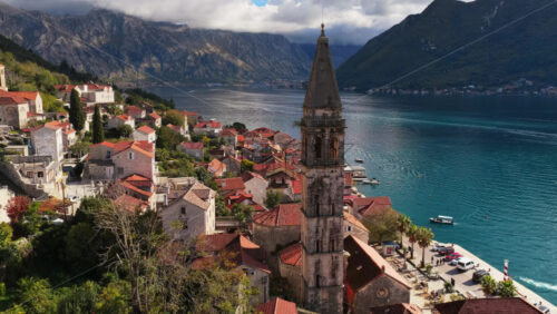 Video - Aerial drone view of the bell tower in Perast rising above traditional stone houses, with the Bay of Kotor and steep mountains visible behind it. Bay of Kotor, Montenegro