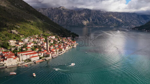 Video - Aerial drone view of the hillside town of Perast, highlighting the tall church tower, dense red roofed houses, and the waterfront along the Bay of Kotor