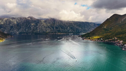 Video - Aerial drone view of the Bay of Kotor, with small boats creating wake trails on calm water and towering rocky mountains under cloudy skies