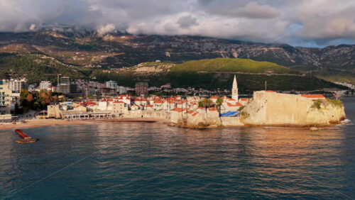 Video - Aerial drone view of Budva Old Town, showing its medieval stone buildings, red rooftops, fortress walls, and church tower along the coastline, with modern city buildings and mountains in the background. Montenegro