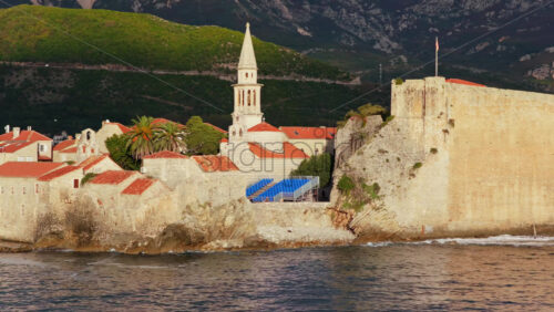 Video - Aerial drone view of Budva Old Town, showing its medieval stone buildings, red rooftops, fortress walls, and church tower along the coastline, with modern city buildings and mountains in the background. Montenegro