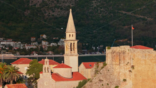 Video - Aerial drone view of Budva Old Town, showing its medieval stone buildings, red rooftops, fortress walls, and church tower along the coastline, with modern city buildings and mountains in the background. Montenegro
