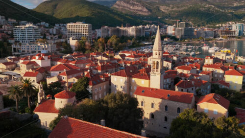 Video - Aerial drone view of Budva Old Town, showing its medieval stone buildings, red rooftops, fortress walls, and church tower along the coastline, with modern city buildings and mountains in the background. Montenegro