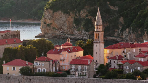 Video - Aerial drone view of Budva Old Town, showing its medieval stone buildings, red rooftops, fortress walls, and church tower along the coastline, with modern city buildings and mountains in the background. Montenegro