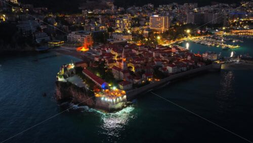 Video - Aerial drone view of Budva after sunset, featuring stone houses with red roofs and a tall church bell tower warmly illuminated from within. Montenegro