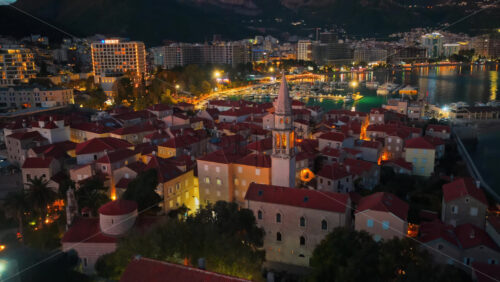 Video - Aerial drone view of Budva after sunset, featuring stone houses with red roofs and a tall church bell tower warmly illuminated from within. Montenegro
