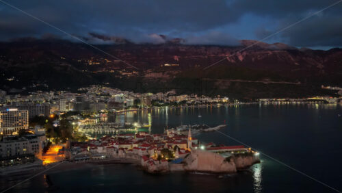 Video - Aerial drone view of Budva after sunset, featuring stone houses with red roofs and a tall church bell tower warmly illuminated from within. Montenegro