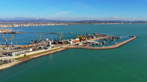 Video - Aerial drone view of a busy container port, showing cargo ships, stacked colorful containers, industrial cranes, and long breakwaters extending into the sea in Durres, Albania