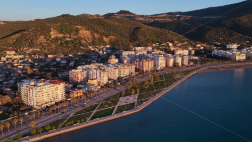 Video - Aerial drone view of Vlora's modern coastline at golden hour, with warm sunlight illuminating residential buildings and lush forested hills in the background