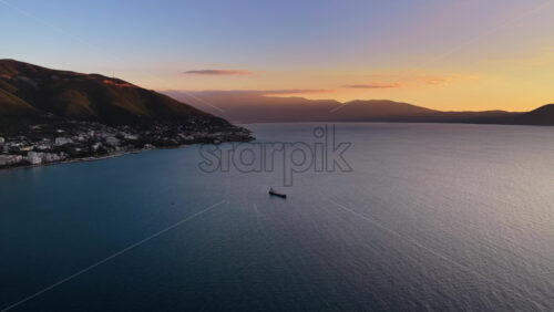 Video - Aerial drone view of a lone fisherman rowing his small wooden boat during sunset in Vlora Bay, Albania, with soft orange skies, calm waters, and distant silhouettes of hills and clouds