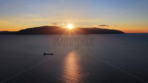 Video - Aerial drone view of a lone fisherman rowing his small wooden boat during sunset in Vlora Bay, Albania, with soft orange skies, calm waters, and distant silhouettes of hills and clouds