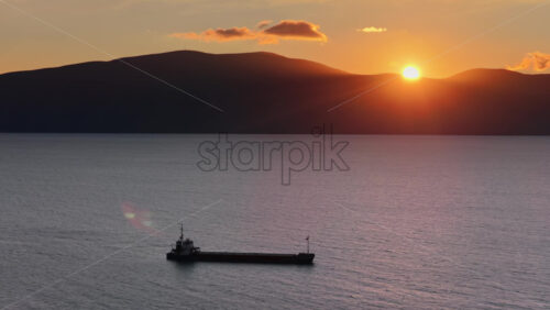 Video - Aerial drone view of a lone fisherman rowing his small wooden boat during sunset in Vlora Bay, Albania, with soft orange skies, calm waters, and distant silhouettes of hills and clouds