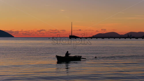 Video - Aerial drone view of a lone fisherman rowing his small wooden boat during sunset in Vlora Bay, Albania, with soft orange skies, calm waters, and distant silhouettes of hills and clouds